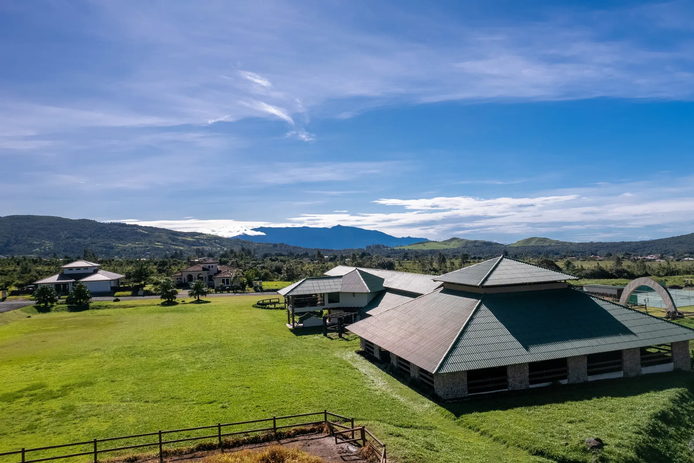 Photo of the Boquete Country Club grounds, with a large green-roofed building on stone pillars beside open lawn, nearby homes, a tennis court and mountains under a blue sky.