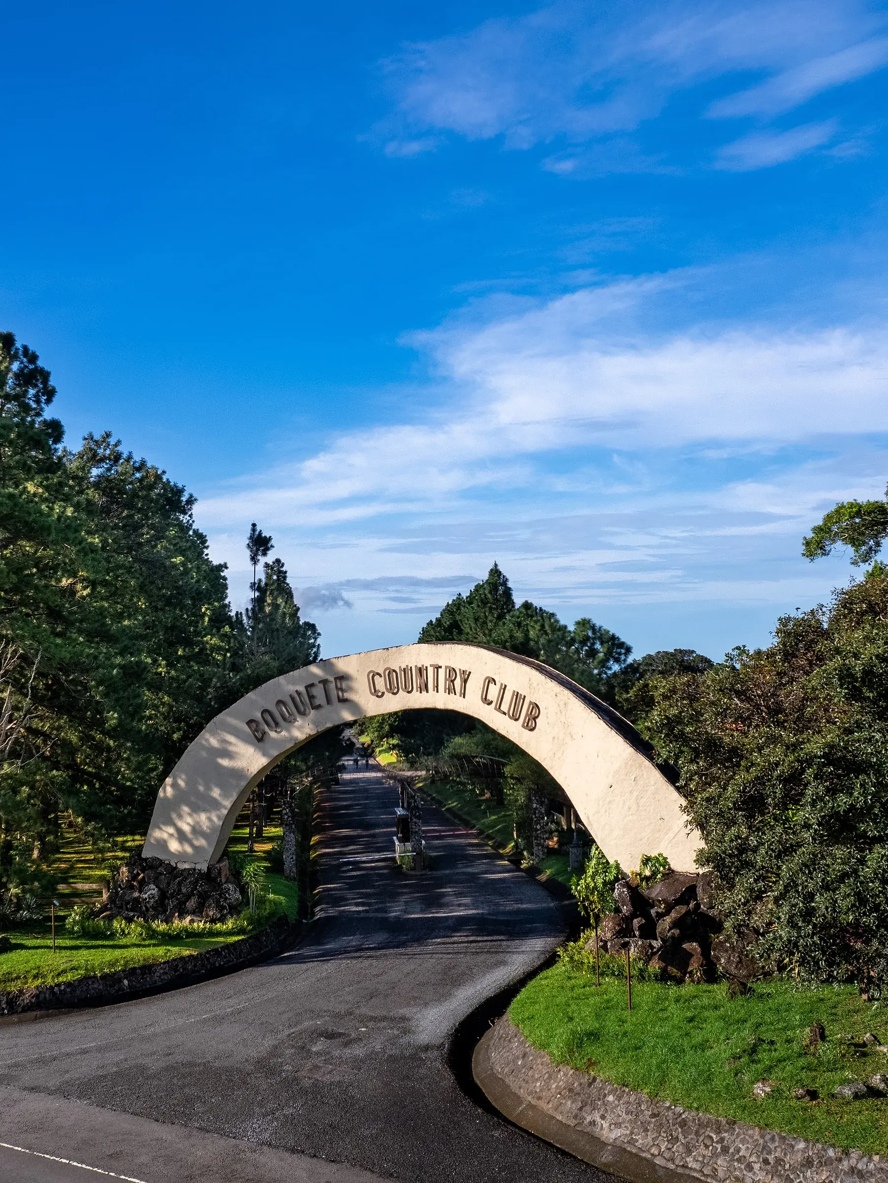 Photo of the Boquete Country Club entrance arch over a paved road lined with pines and stone-edged lawns, with the club name on the arch.