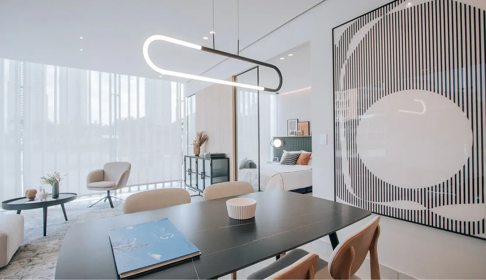 Photo of a furnished Generation Tower suite, with a dark dining table and a pendant light in the foreground, a living area on the left and the bedroom behind a glass partition.