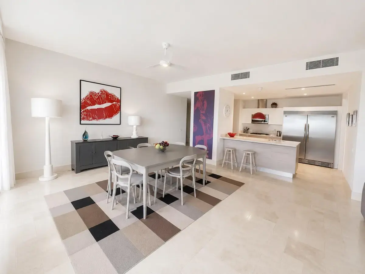 Comedor en Puntarena Ocean Village con mesa gris para seis sobre una alfombra a cuadros y, al fondo, la cocina abierta con refrigeradora doble de acero.