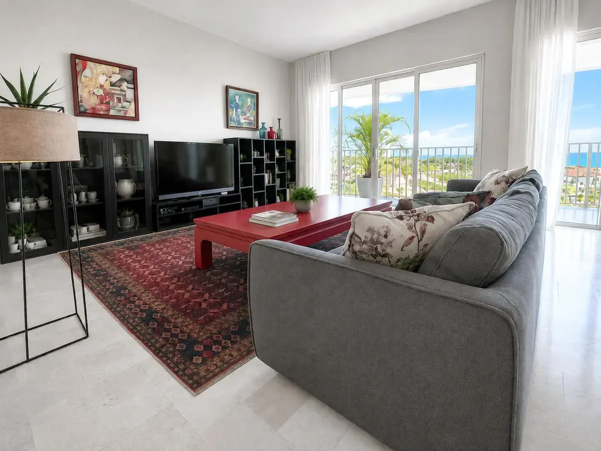 Sala en Puntarena Ocean Village con sofá gris, mesa de centro roja lacada, alfombra roja, repisas negras con televisor y puertas de vidrio hacia la vista al mar.