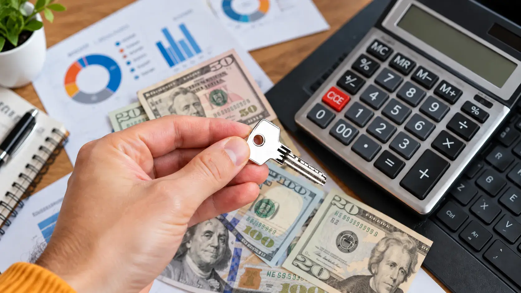 A hand holds a house key over US dollar bills, with a calculator and printed charts on the desk