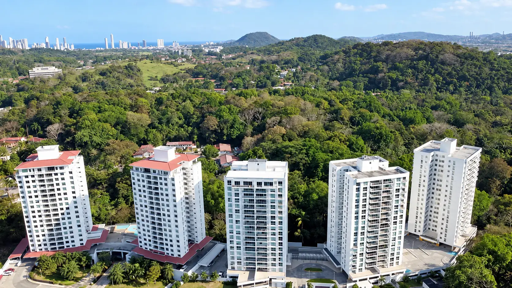 Vista aérea de las torres blancas de Clayton Park II sobre el dosel de árboles, con el horizonte de la Ciudad de Panamá y la costa del Pacífico al fondo