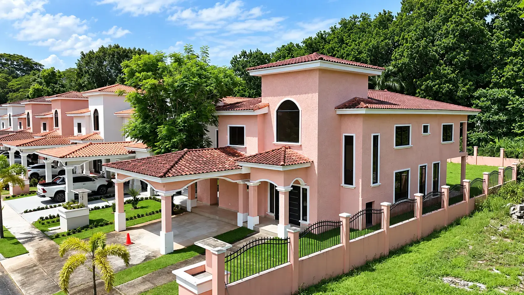 A two-story pink stucco house with a terracotta tile roof and covered carport, part of a row of similar homes behind a black metal perimeter fence