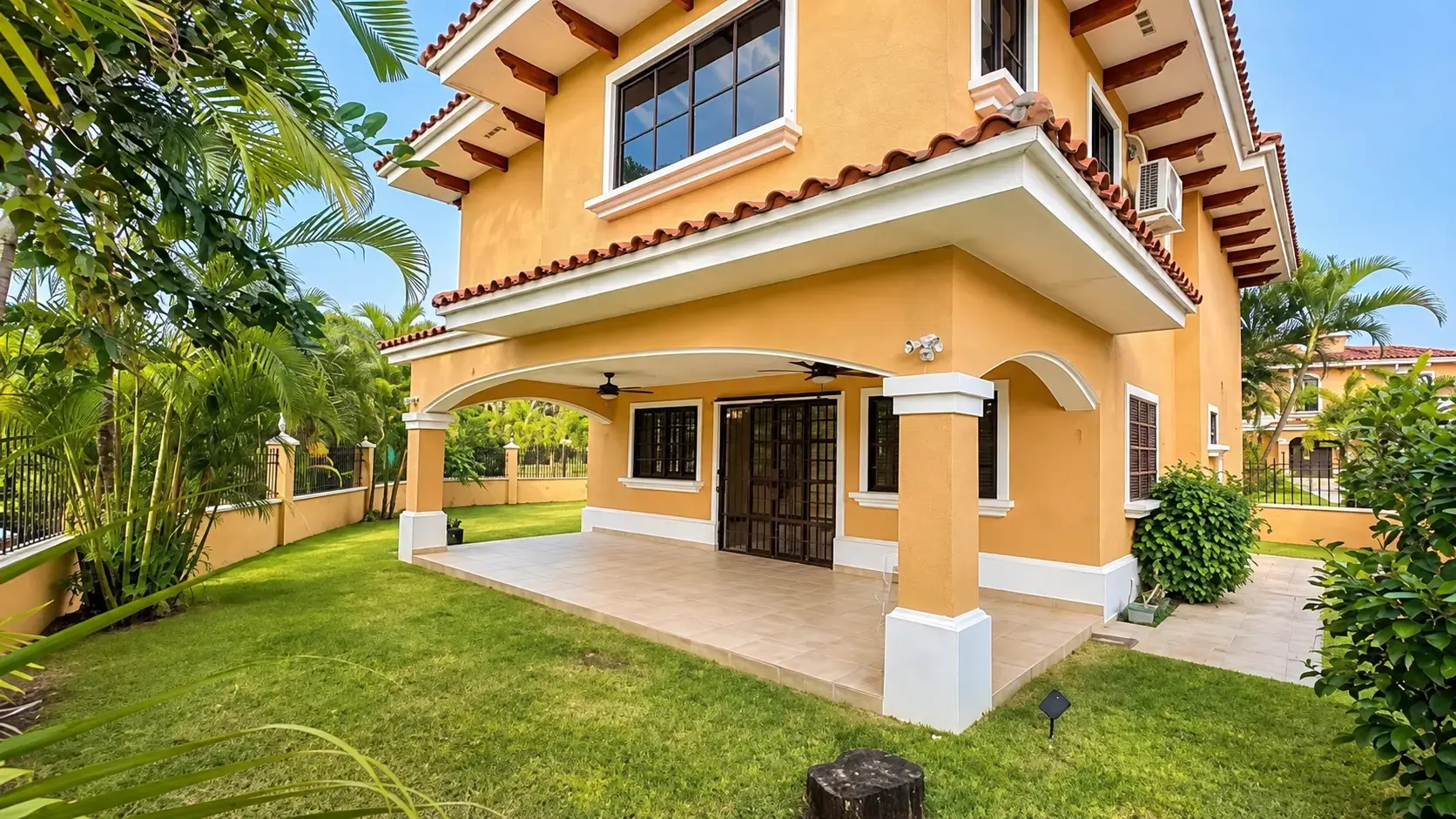 A two-story yellow stucco house with a covered rear patio, a ceiling fan, and tropical garden landscaping