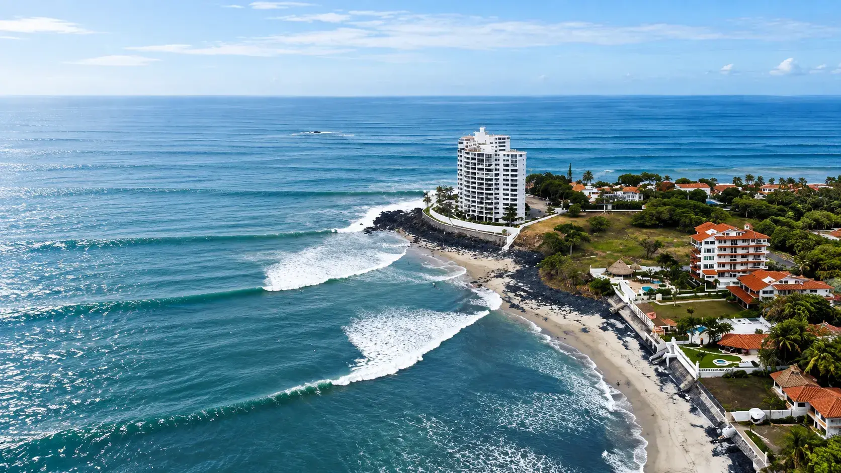 Vista aérea de una punta rocosa en la costa del Pacífico, con olas que entran a una bahía de arena y edificios bajos y jardines junto al mar