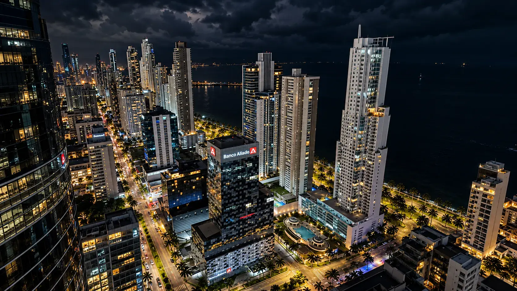 Vista aérea nocturna de torres residenciales y de oficinas iluminadas sobre una avenida costera con palmeras, con la bahía de Panamá oscura al fondo